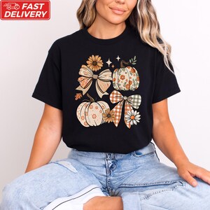 A woman wearing a black t-shirt with a graphic design of pumpkins and bows, sitting on the floor.