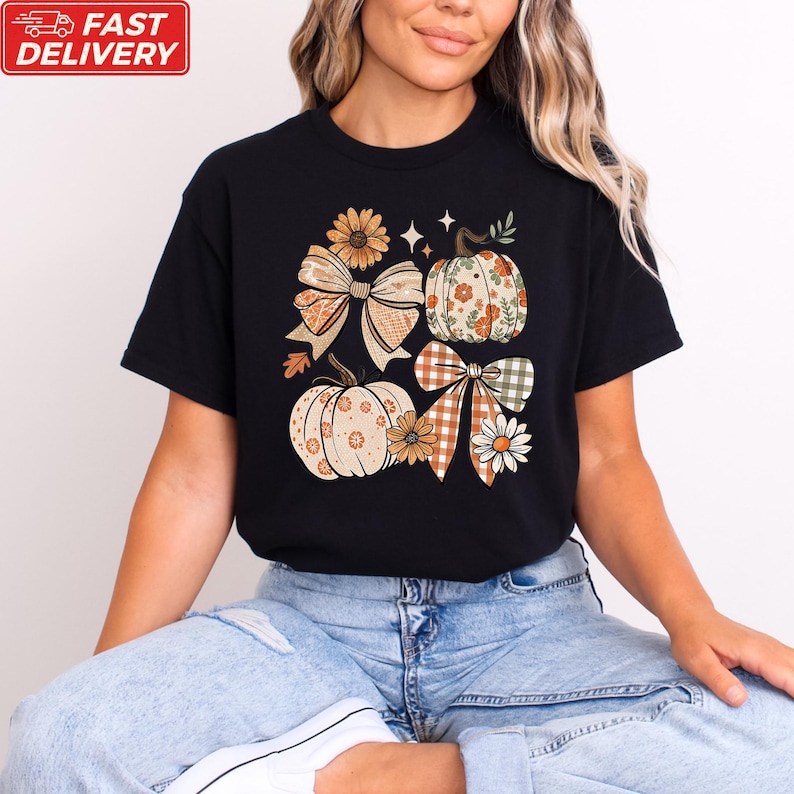 A woman wearing a black t-shirt with a graphic design of pumpkins and bows, sitting on the floor.