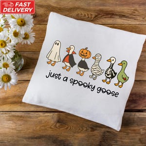 A white pillow with a Halloween-themed design featuring cartoon goose characters, including a ghost, pumpkin, mummy, skeleton, and a green goose.