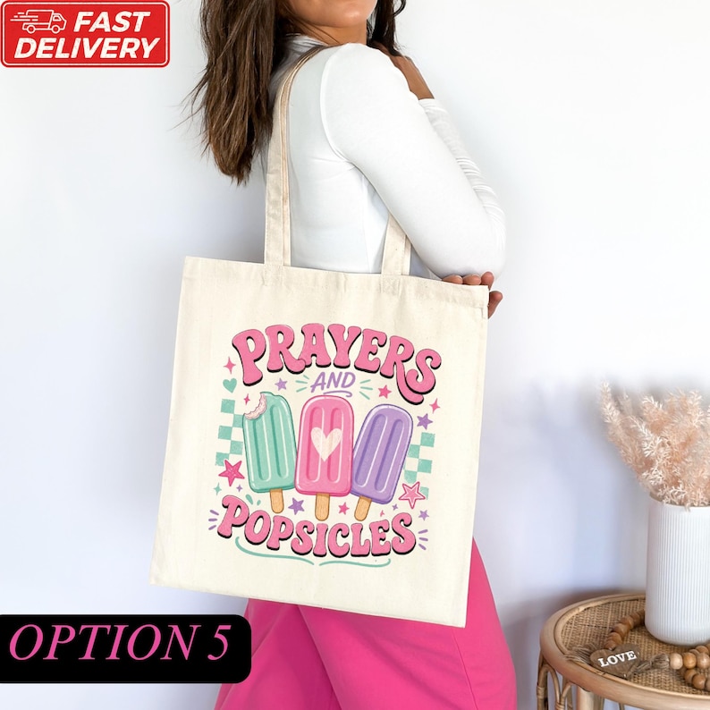 A woman is holding a white tote bag with a colorful graphic design featuring three popsicles and the text "Prayers and Popsicles".