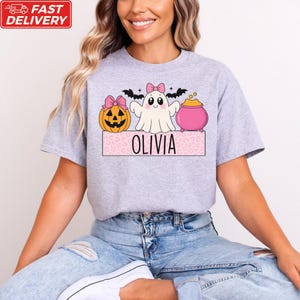A woman wearing a gray t-shirt with a Halloween-themed graphic design featuring a ghost, pumpkin, and a pot of gold.
