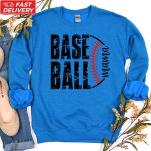 A blue sweatshirt with the text "BASE BALL" printed on it, along with a baseball graphic. The sweatshirt is placed on a white background, and there are some flowers and a pair of shoes visible in the image.