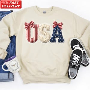A beige sweater with the word "USA" embroidered on it, along with a pair of black and white sneakers, a white mug, and a pair of blue jeans.