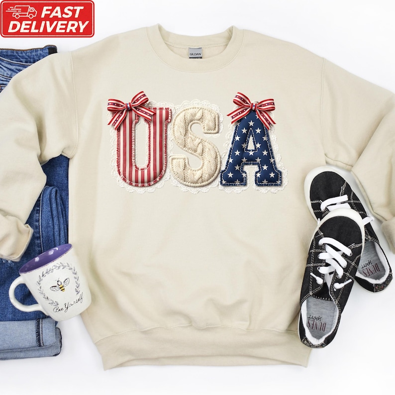 A beige sweater with the word "USA" embroidered on it, along with a pair of black and white sneakers, a white mug, and a pair of blue jeans.