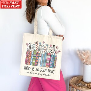 A woman is standing next to a table with a white vase on it, wearing a white shirt and pink pants. She is carrying a white tote bag with a colorful graphic design and text printed on it.