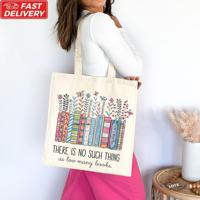A woman is standing next to a table with a white vase on it, wearing a white shirt and pink pants. She is carrying a white tote bag with a colorful graphic design and text printed on it.