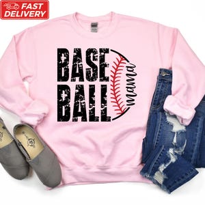 A pink sweatshirt with the text "BASE BALL" printed on it, along with a baseball graphic and the word "Momma" below it. The sweatshirt is placed on top of a pair of jeans and a pair of gray shoes.