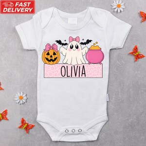 A white baby onesie with a Halloween-themed design, including a ghost, pumpkin, and a pot of gold. The name "OLIVIA" is written on the onesie.