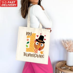 A woman is holding a white tote bag with a printed design of a turkey wearing a hat and the text "My 1st Thanksgiving".