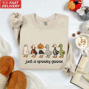 a beige sweatshirt with a Halloween-themed graphic design, including a skeleton, mummy, and ghost, along with the text "Just a spooky goose". The sweatshirt is laid out on a white surface, surrounded by various Halloween-themed items such as a pumpkin, a cup of hot chocolate, and a book.