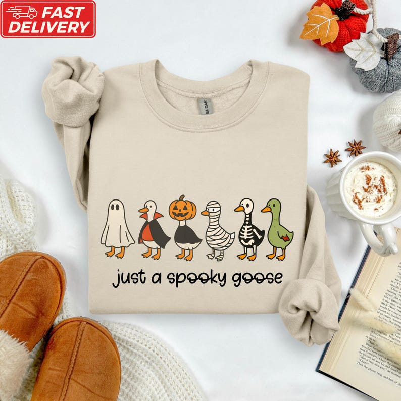 a beige sweatshirt with a Halloween-themed graphic design, including a skeleton, mummy, and ghost, along with the text "Just a spooky goose". The sweatshirt is laid out on a white surface, surrounded by various Halloween-themed items such as a pumpkin, a cup of hot chocolate, and a book.