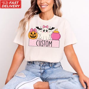 A woman wearing a white t-shirt with a Halloween-themed graphic design, including a ghost, pumpkin, and a pink pot.