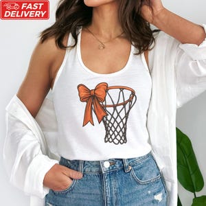 A woman wearing a white tank top with an orange bow on it, paired with blue jeans, stands in front of a plant.