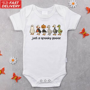 A white baby onesie with a Halloween-themed graphic of a ghost, pumpkin, skeleton, and geese, along with the text "Just a spooky goose".