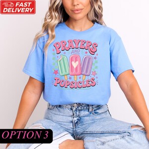 A woman wearing a blue t-shirt with a graphic design of three popsicles and the text "Prayers and Popsicles" printed on it.