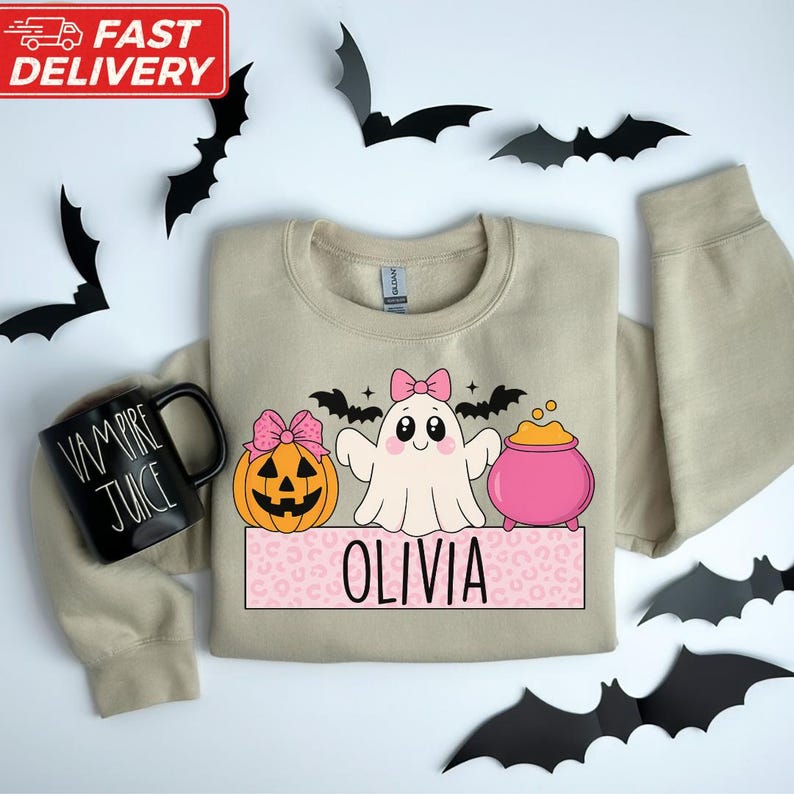 a beige sweatshirt with a Halloween-themed design, including a ghost, pumpkin, and a mug with the text "VA VAPOR JUICE". The sweatshirt is surrounded by black bats, and the word "OLIVIA" is displayed below the design.