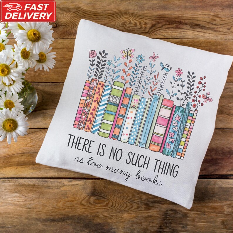 A white pillow with a colorful illustration of books and flowers, accompanied by the text "THERE IS NO SUCH THING as too many books" on a wooden surface.