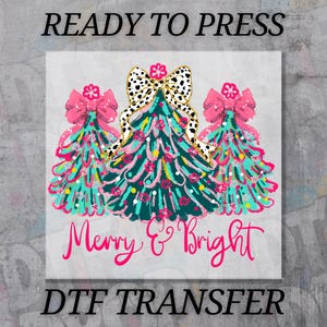May include: A festive design featuring three Christmas trees with pink bows and a dalmatian print bow. The trees are teal and pink with colorful accents. The words "Merry & Bright" are written in pink script. The text "READY TO PRESS" and "DTF TRANSFER" are also present.