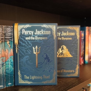May include: Two hardcover books from the "Percy Jackson and the Olympians" series on a bookshelf. The first book, "The Lightning Thief," is blue with gold accents and a trident illustration. The second book is "Sea of Monsters."