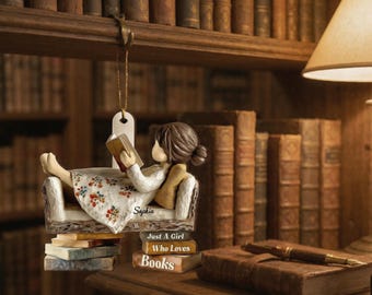May include: A decorative ornament of a girl reading a book while reclining on a small chair. The ornament is suspended in front of a bookshelf. The text on the ornament reads "Just a Girl Who Loves Books".