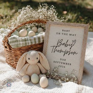 May include: A wicker basket filled with eggs and a knitted bunny toy. A letter board with the text "Baby on the Way" and "Baby Thompson, Due September 2026" is displayed. The scene is decorated with white flowers.