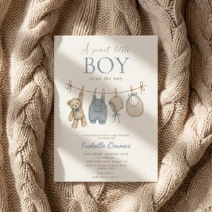 Peut inclure: Une carte d'invitation pour baby shower de couleur crème avec le texte A sweet little BOY is on the way. Le design présente une illustration d'un ours en peluche, d'une salopette bleue, d'un bonnet et d'un bavoir suspendus à une corde à linge.