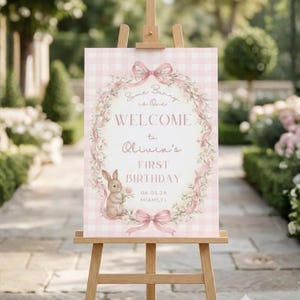 Pode incluir: Uma placa de boas-vindas rosa e branca xadrez em um cavalete de madeira. A placa apresenta uma coroa de flores, uma ilustração de coelho e o texto "Welcome to Olivia's First Birthday" com a data e o local.