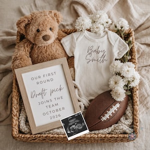 Puede incluir: Una cesta de mimbre contiene un oso de peluche, un body blanco con "Baby Smith" impreso, un balón de fútbol americano y un anuncio enmarcado que dice "Our First Round Draft Pick Joins The Team October 2026". Flores blancas dispuestas alrededor.
