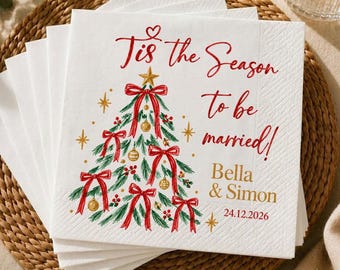 May include: A stack of white paper napkins featuring a green Christmas tree illustration with red bows and gold ornaments. The text reads 'Tis the Season to be married!', 'Bella & Simon', and the date '24.12.2026' in red and gold script.