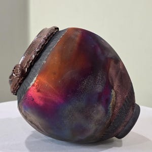 May include: A handcrafted ceramic vase with a unique, iridescent finish. The vase features a dark gray rim and base, with a body that transitions from orange to purple hues. The textured surface adds depth to the design. The vase is approximately 8 inches tall.