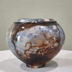 May include: A round ceramic bowl with a wide opening and a short, narrow base. The surface features a mottled glaze with blended tones of dark brown, charcoal, and pale blue, creating a textured, smoky appearance across the rounded body.