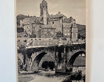 May include: Black and white vintage travel poster of Estaing, France, featuring a stone bridge and castle. The word "FRANCE" is prominently displayed at the bottom. The poster is held by two black binder clips.