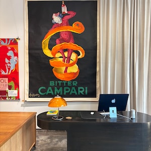 May include: A vintage Campari poster with a figure in a red polka-dot outfit holding a bottle, set against an orange peel design. The poster hangs above a dark desk with a laptop and an orange lamp.