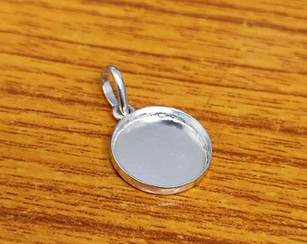 May include: A small, round silver pendant with a loop at the top. The pendant has a recessed area, likely for setting a stone or other decorative element. The pendant is shown on a wooden surface.