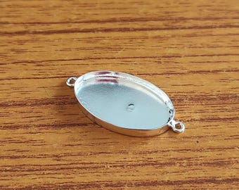 May include: A silver-colored oval bezel setting with two small loops for attaching to other components. The setting has a shallow, recessed area for holding a cabochon or other decorative element. The setting is on a wooden surface.
