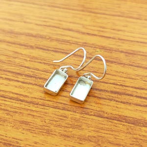 May include: A pair of silver-toned rectangular earrings with a hook design. The earrings have a rectangular frame with a light-colored interior. The earrings are displayed on a wooden surface.