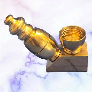 May include: A gold-colored pipe with a cylindrical body and a bowl-shaped head. The pipe is made of metal and has a decorative design on the mouthpiece. It sits on a small, angled base.