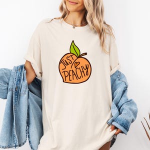 May include: Cream-colored t-shirt featuring a cartoon peach with the words "Just Peachy" in black and orange. The peach has a green leaf and a small heart. The shirt is a relaxed fit.