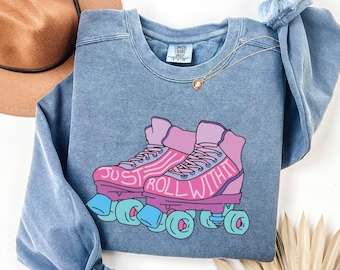 May include: A blue sweatshirt featuring a graphic of pink roller skates with the words "JUST ROLL WITH IT". The skates have blue wheels and laces. A gold necklace and a blue scrunchie are also visible.