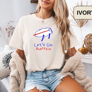 May include: Ivory-colored t-shirt with a blue and red graphic of a buffalo and the text "Let's Go Buffalo." The shirt is a relaxed fit and is paired with denim shorts. The word "IVORY" is in a cream-colored rectangle.