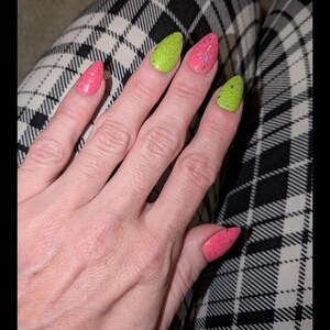 Peut inclure: Une main avec de longs ongles pointus peints de couleurs vives. Les ongles sont roses et vert néon, avec des paillettes. L'arrière-plan est un motif à carreaux noir et blanc.
