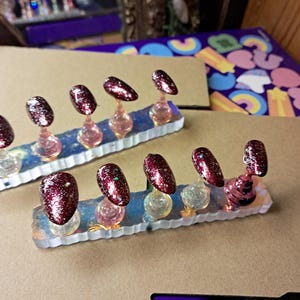Peut inclure: Ensemble de faux ongles avec une finition pailletée bordeaux. Chaque ongle est monté sur un support transparent avec une base rose ou transparente. Les ongles sont disposés sur une base rectangulaire irisée.