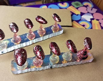 May include: Set of artificial nails with a burgundy glitter finish. Each nail is mounted on a clear, decorative stand with a pink or clear base. The nails are arranged on a rectangular, iridescent base.
