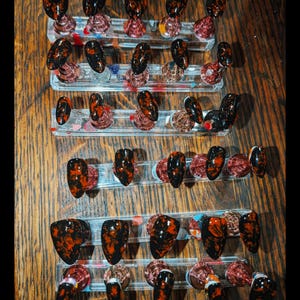 May include: A collection of press-on nails arranged on clear acrylic stands. Each nail features a glossy black base with a marbled pattern of vibrant red and orange streaks. The set includes various sizes and shapes, all displaying a consistent dark, fiery color scheme across the entire display of individual nail pieces.