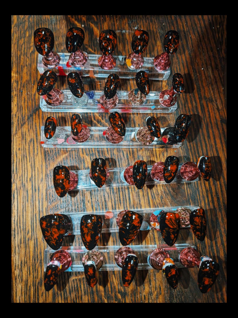 May include: A collection of press-on nails arranged on clear acrylic stands. Each nail features a glossy black base with a marbled pattern of vibrant red and orange streaks. The set includes various sizes and shapes, all displaying a consistent dark, fiery color scheme across the entire display of individual nail pieces.