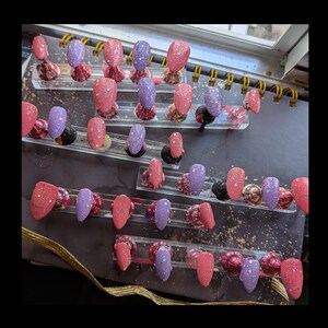 May include: An assortment of artificial nails in shades of pink and purple, some with glitter. The nails are arranged on clear display stands, highlighting diverse designs and textures. The overall look is glamorous and feminine.