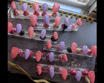 May include: An assortment of artificial nails in shades of pink and purple, some with glitter. The nails are arranged on clear display stands, highlighting diverse designs and textures. The overall look is glamorous and feminine.