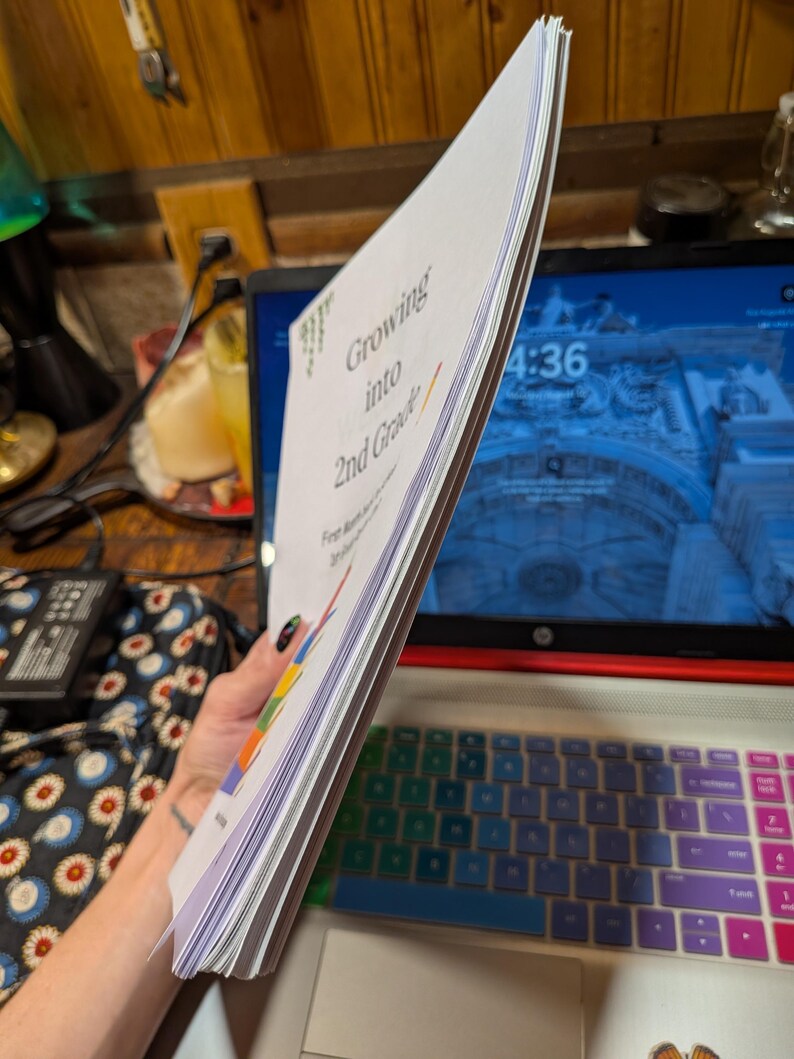 Peut inclure: Une pile de feuilles de papier blanc reli&eacute;es ensemble, avec le texte imprim&eacute; "Growing into 2nd Grade" sur la couverture. Le document comprend des &eacute;l&eacute;ments graphiques color&eacute;s et est tenu verticalement, montrant la tranche du papier.