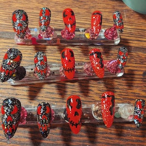 May include: A set of fifteen almond-shaped press-on nails arranged on three clear display stands. The nails feature a mix of red and black base colors with glitter accents and hand-painted black skull face designs with hollow eyes and stitched mouths.