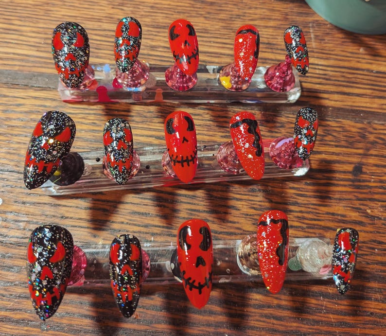 May include: A set of fifteen almond-shaped press-on nails arranged on three clear display stands. The nails feature a mix of red and black base colors with glitter accents and hand-painted black skull face designs with hollow eyes and stitched mouths.